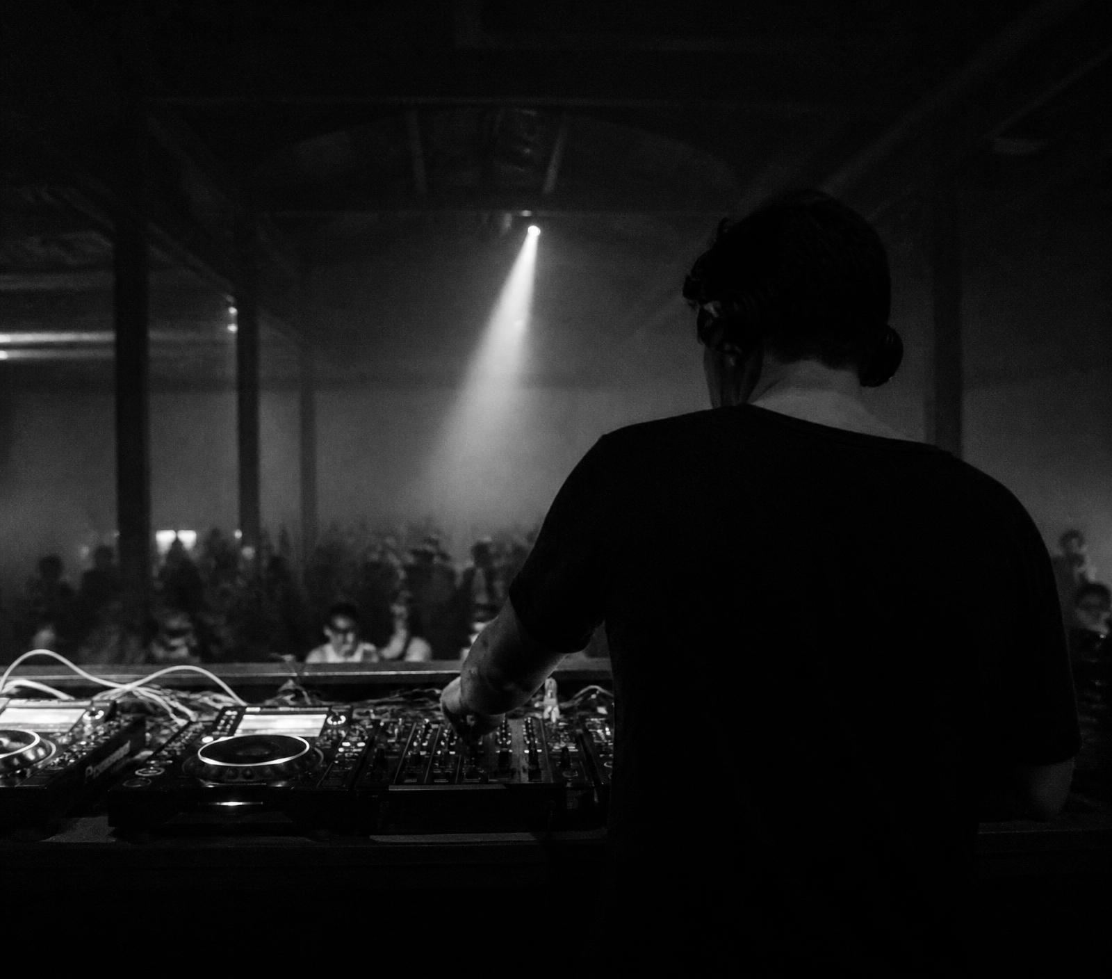 DJ behind the booth in a dark warehouse, single beam of light through haze